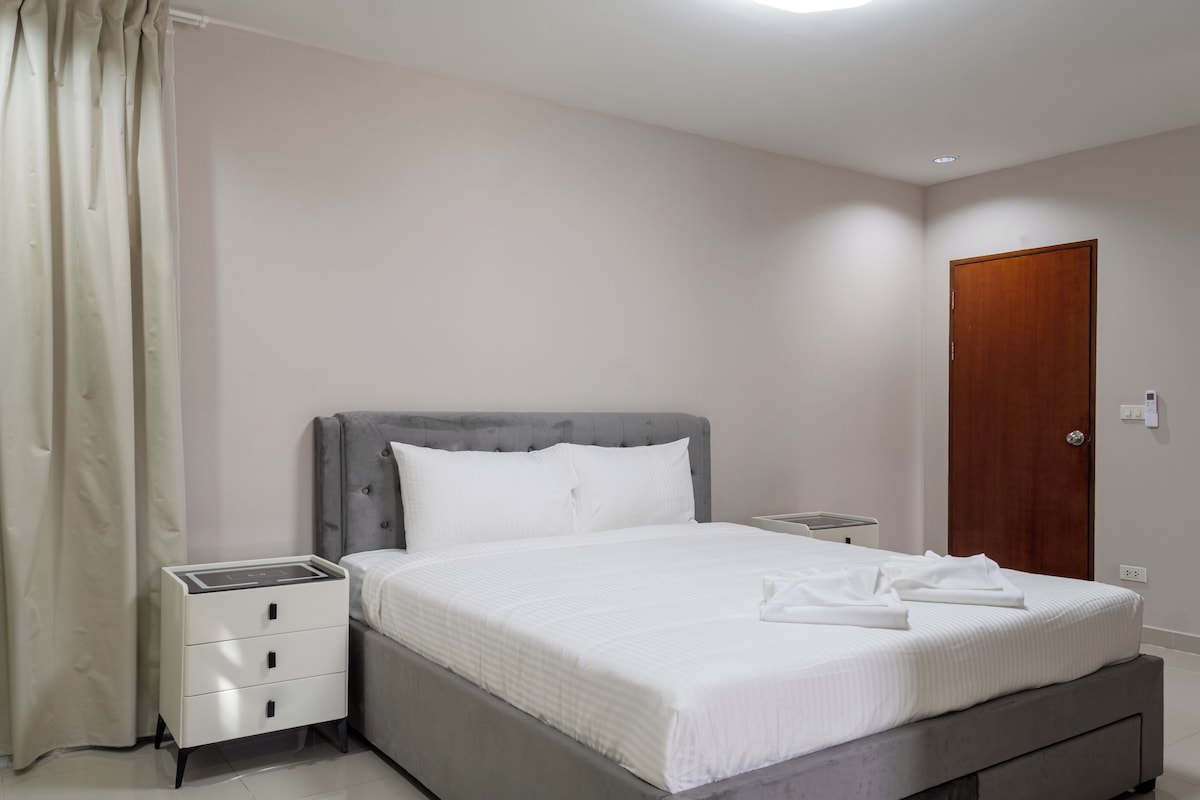A neatly made bed with a grey headboard, a bedside table with a lamp, and a wooden door.