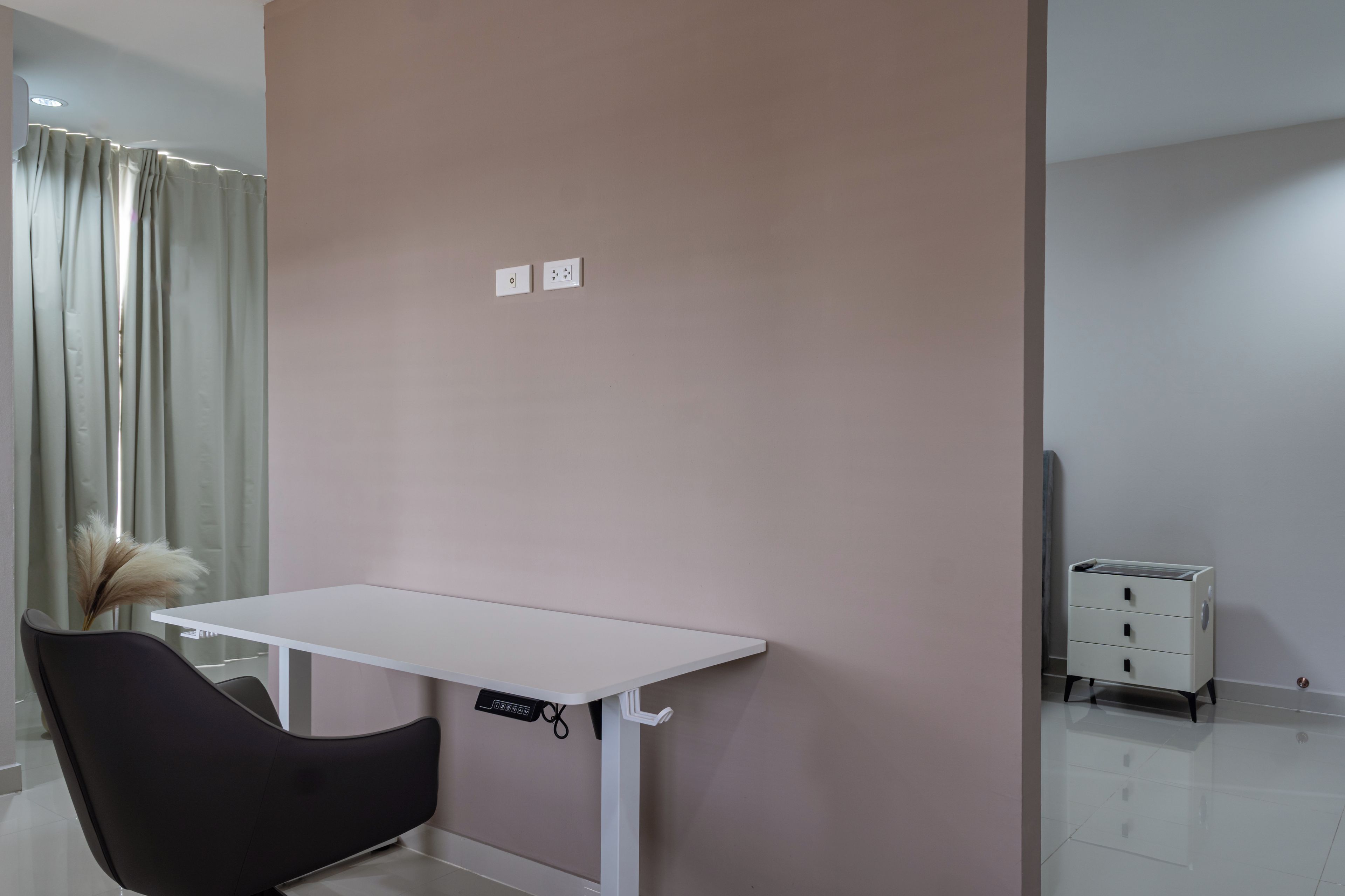 Ein Zimmer mit einem weißen Schreibtisch, einem schwarzen Stuhl, einer rosafarbenen Wand und einem weißen Schrank.