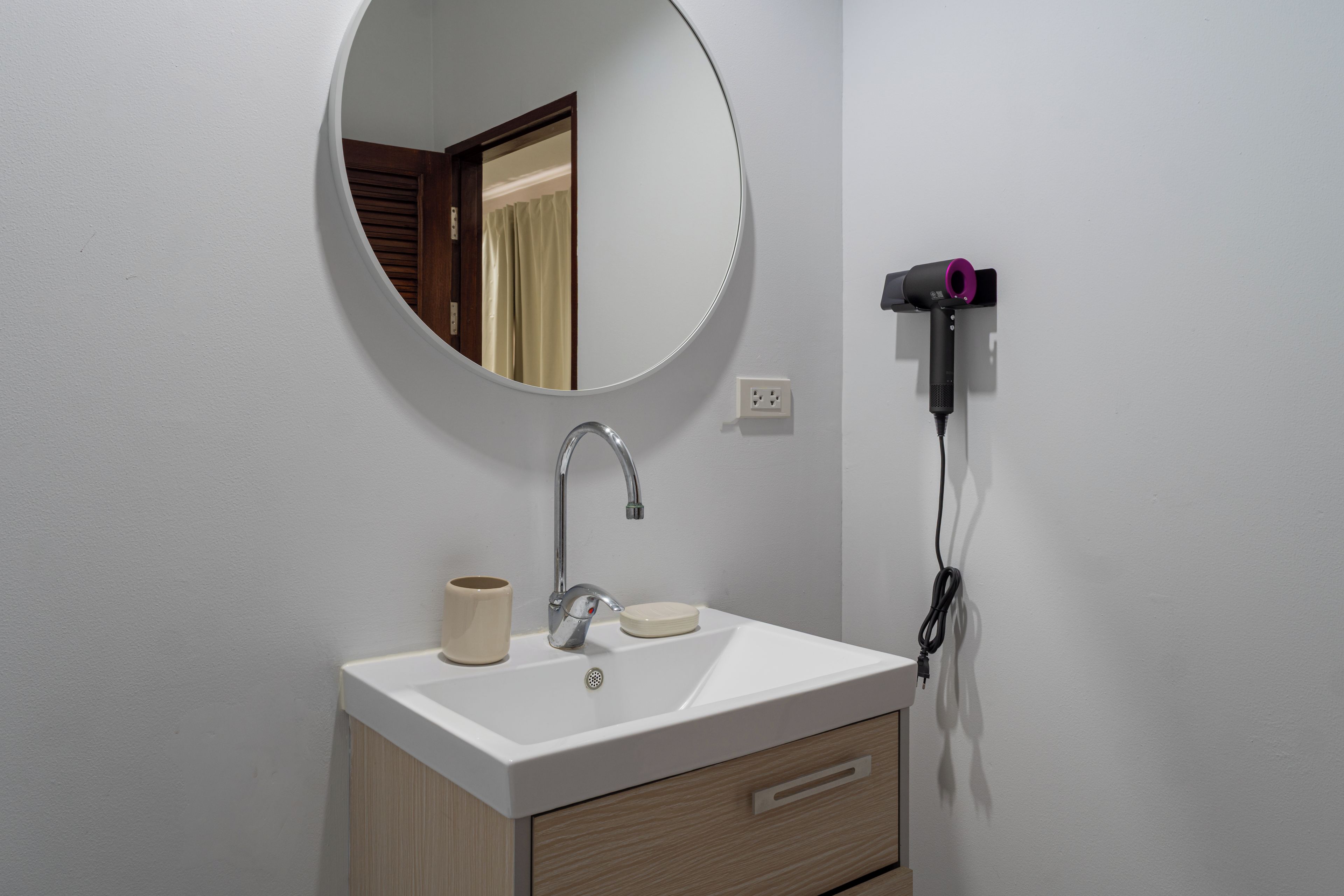 A bathroom with a white sink, a round mirror, a faucet, a cup, and a hairdryer plugged into the wall.