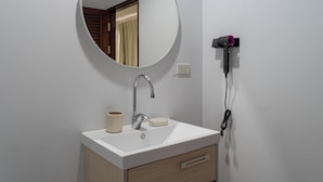 A bathroom with a white sink, a round mirror, a faucet, a cup, and a hairdryer plugged into the wall. - The One Sky  (Rawai)