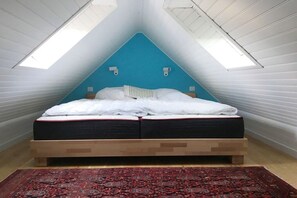 Ein Schlafzimmer mit schräg zulaufender Decke, einem Bett mit weißer Bettwäsche, einem roten Teppich mit Muster und zwei Wandleuchten.