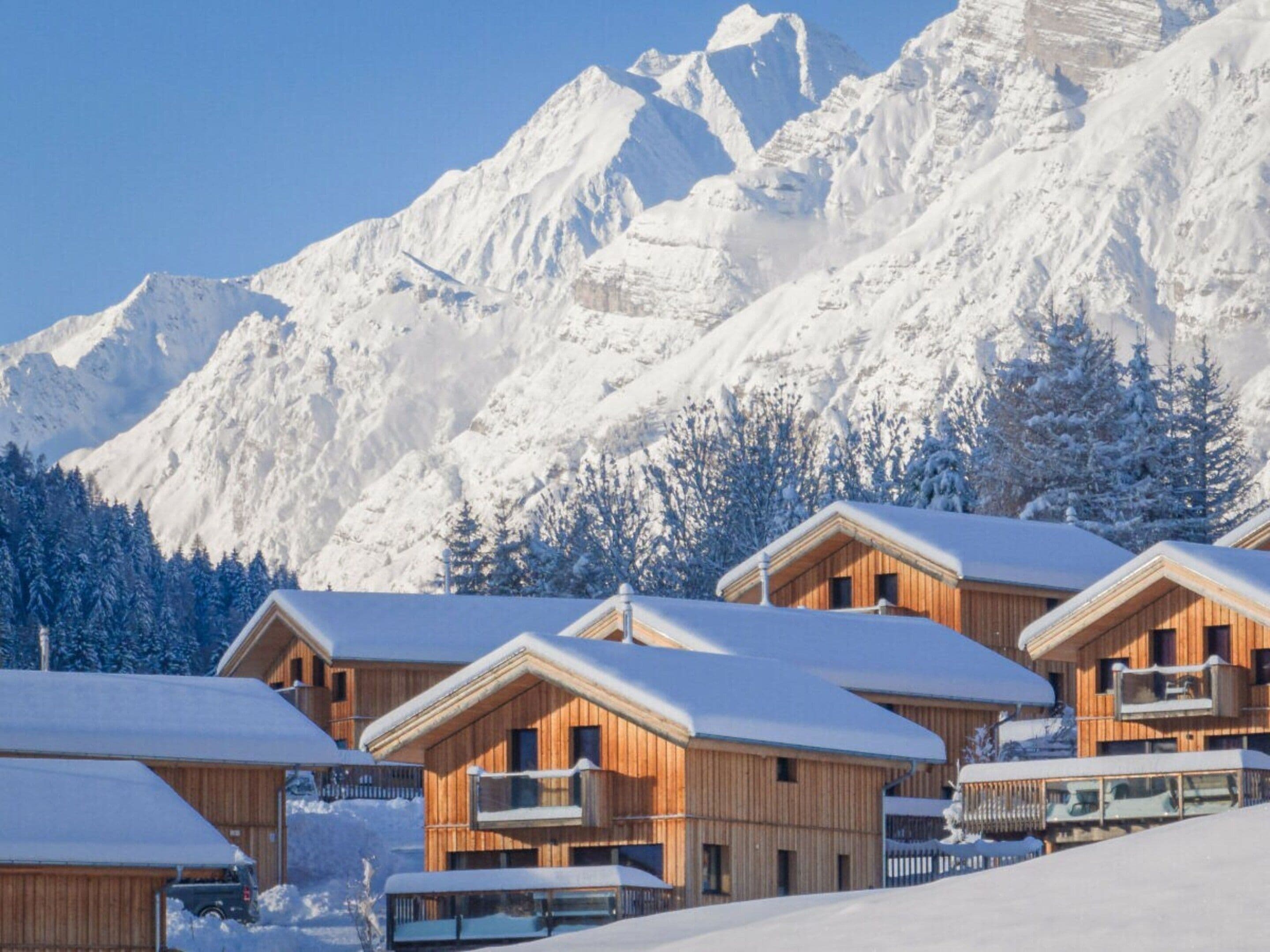 Una hilera de cabañas de madera con techos cubiertos de nieve, frente a un paisaje de montañas nevadas.
