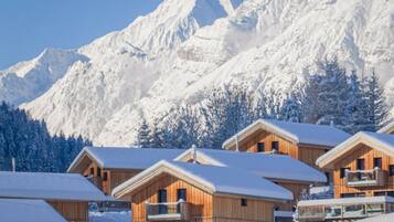 Una hilera de cabañas de madera con techos cubiertos de nieve, frente a un paisaje de montañas nevadas.