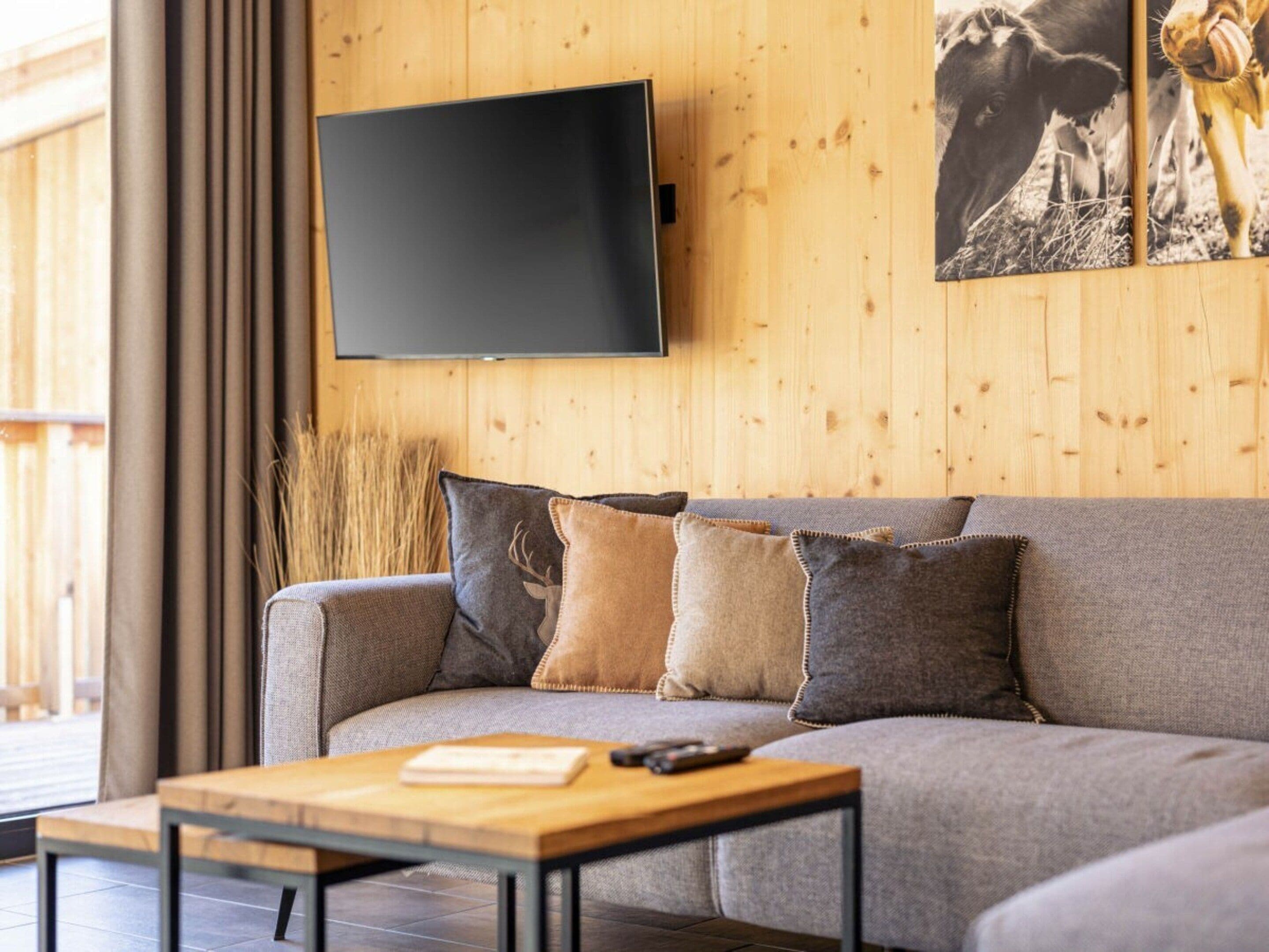 A living room with a grey sofa, a wooden coffee table, a mounted flat-screen TV, and a large window with curtains.