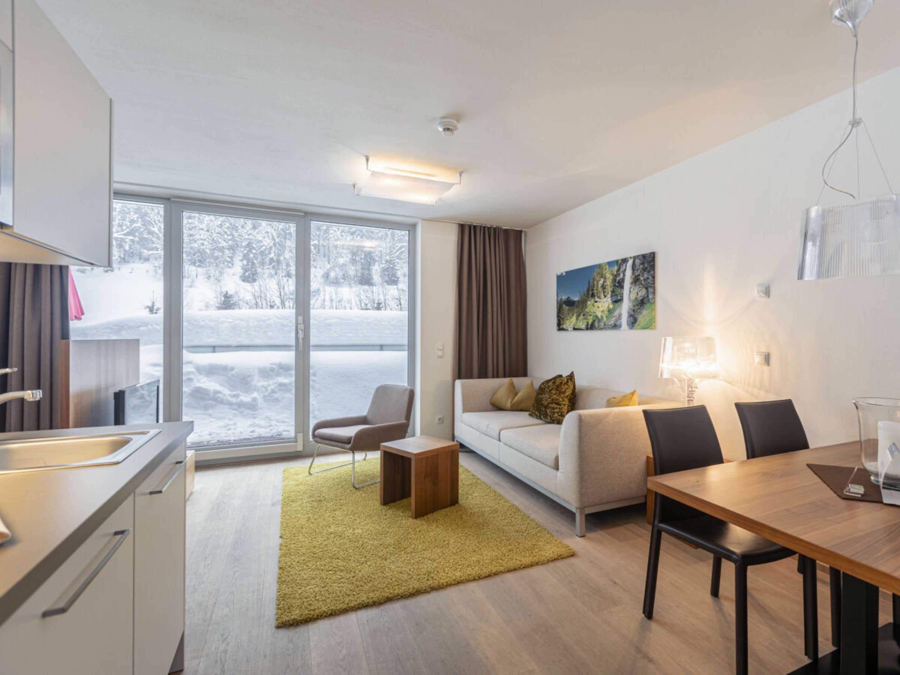 Un salon moderne avec un canapé, une table basse et une table à manger. Une grande fenêtre offre une vue sur un paysage enneigé.