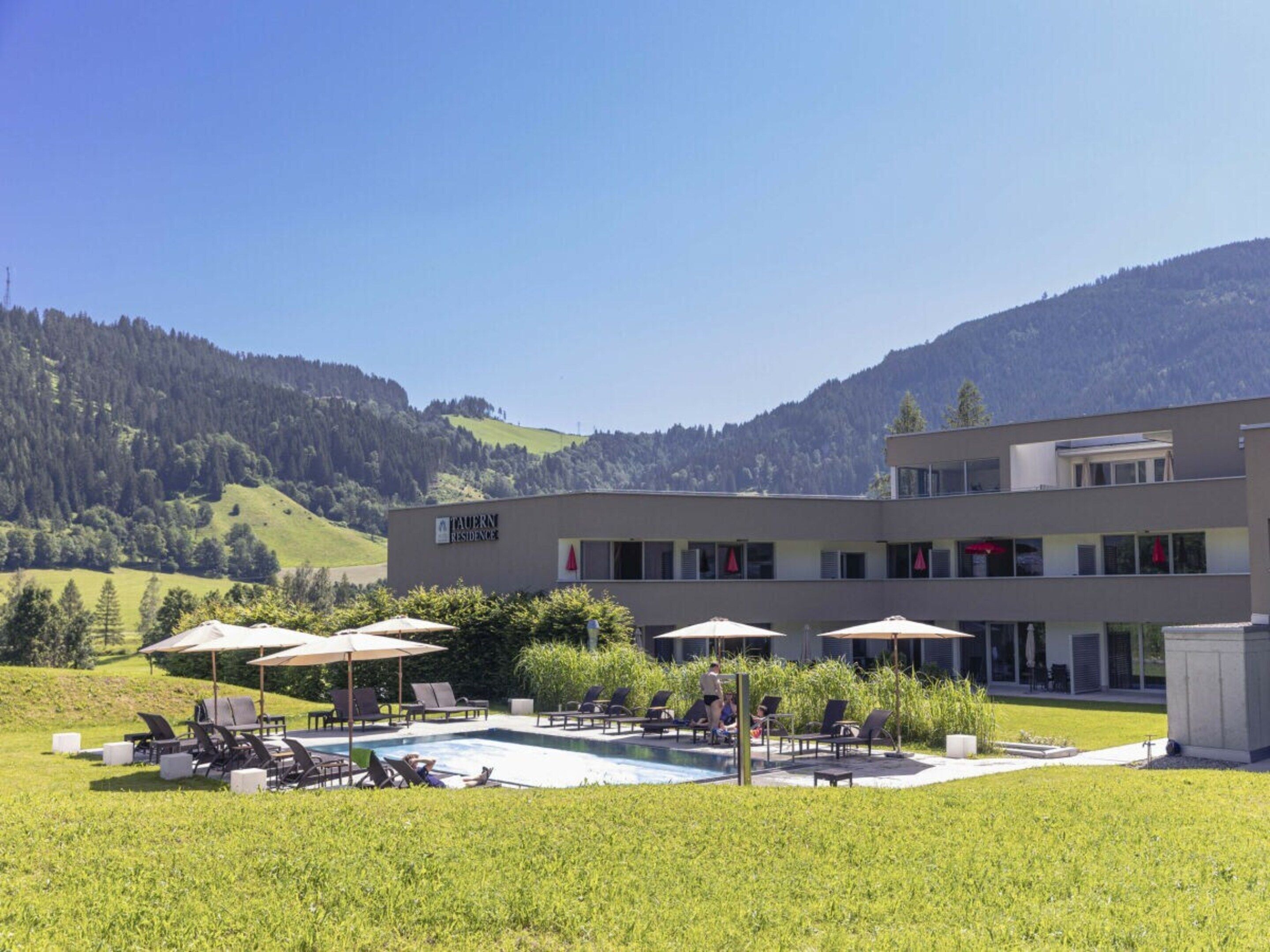 Um edifício moderno com área de piscina, espaço para sentar ao ar livre e vista para uma montanha arborizada.