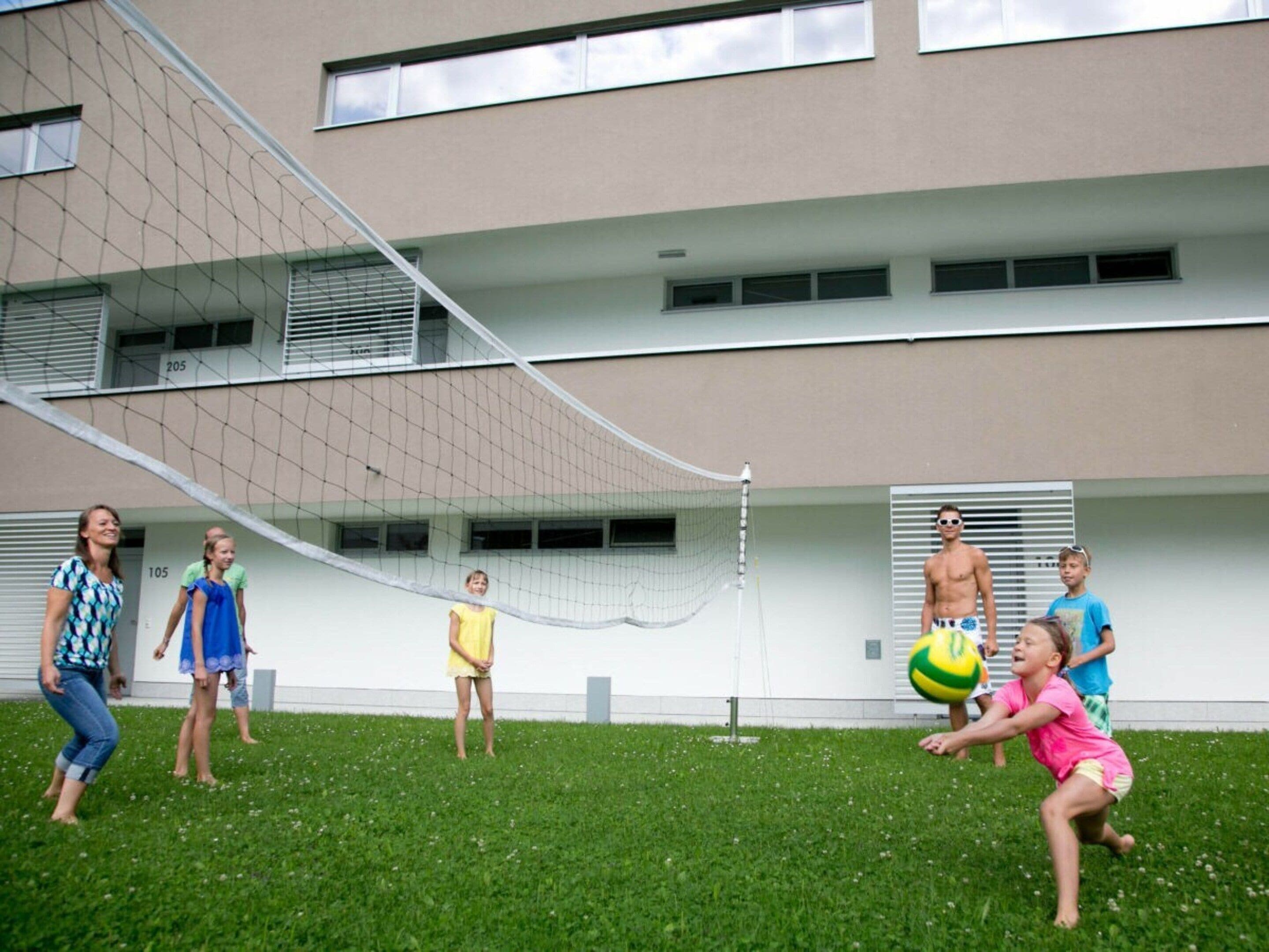 Un gruppo di persone gioca a pallavolo in una zona erbosa, con un edificio sullo sfondo.