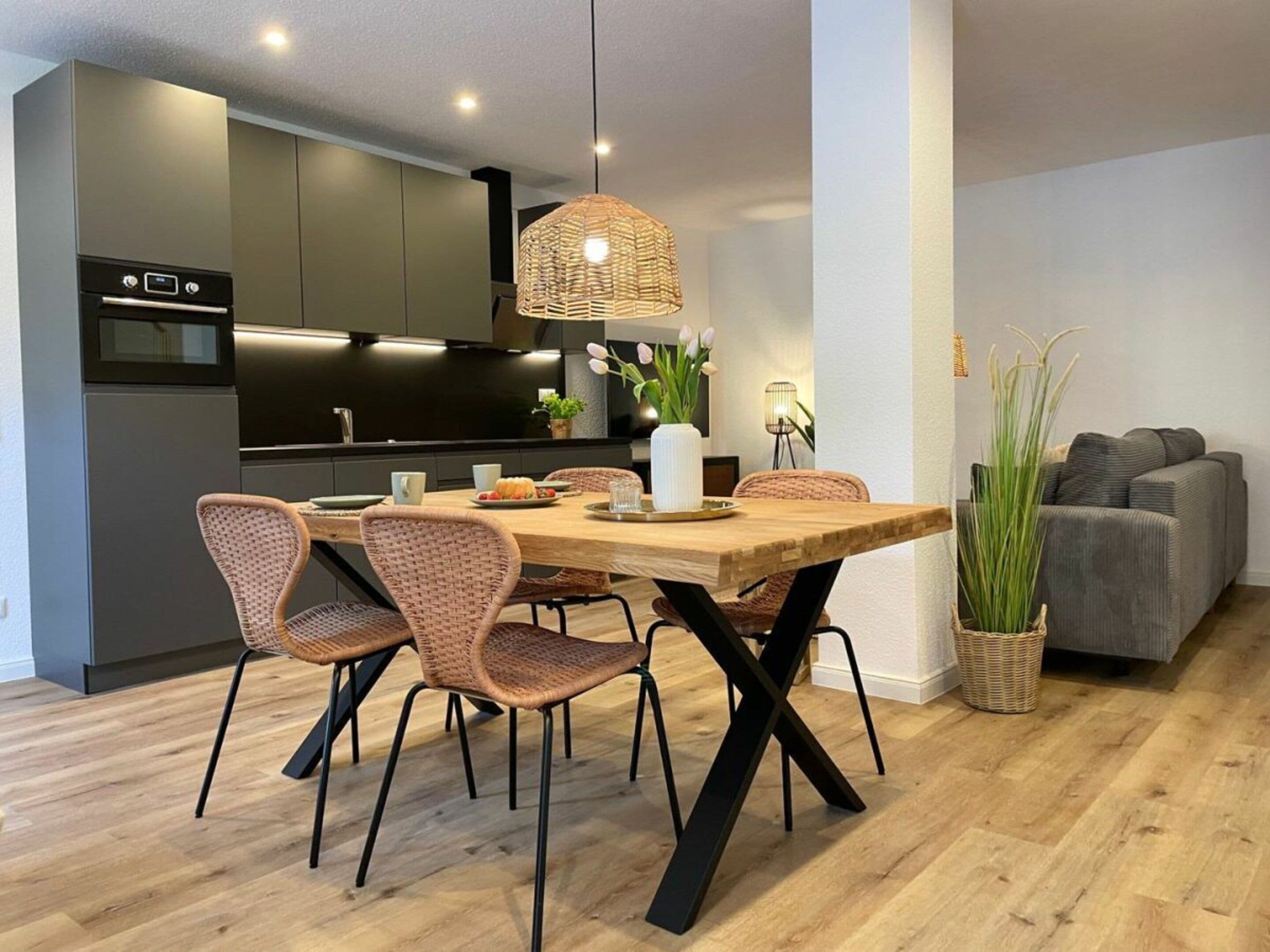 A modern kitchen with dark cabinets, a wooden dining table, wicker chairs, and a hanging light fixture.