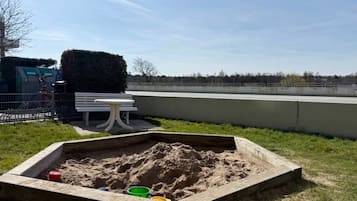 Zona arenosa para jugar con un banco y una mesa, rodeada por una valla y un seto.