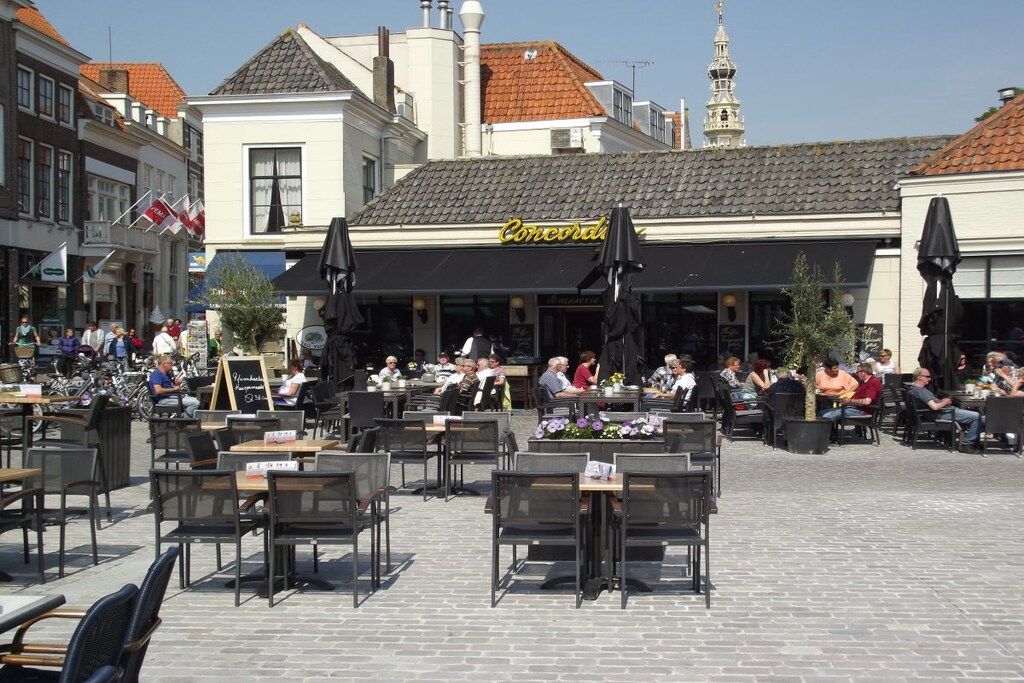 An outdoor café with tables and chairs, a signboard, and a building with a tiled roof in the background.