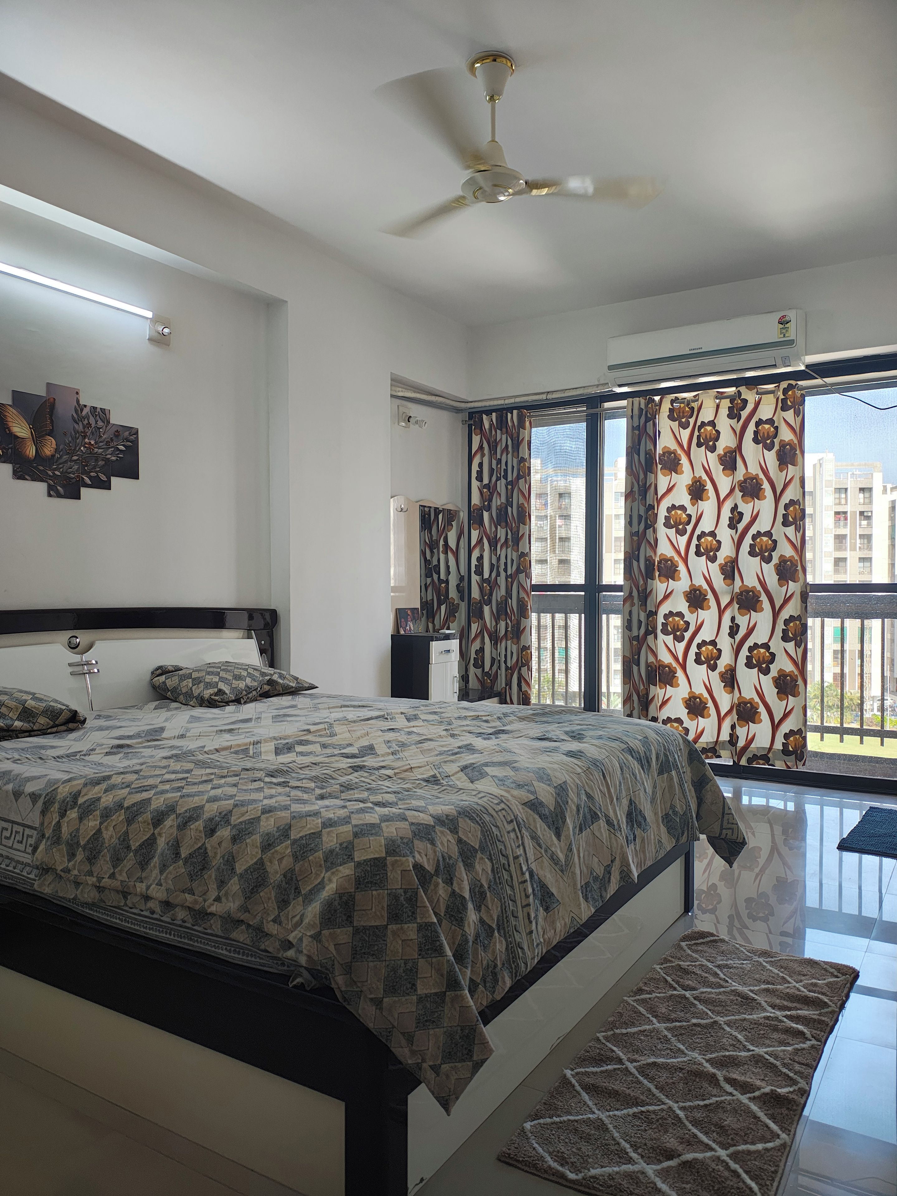 Une chambre à coucher comprenant un lit, un ventilateur de plafond, une fenêtre avec des rideaux et un climatiseur fixé au mur.