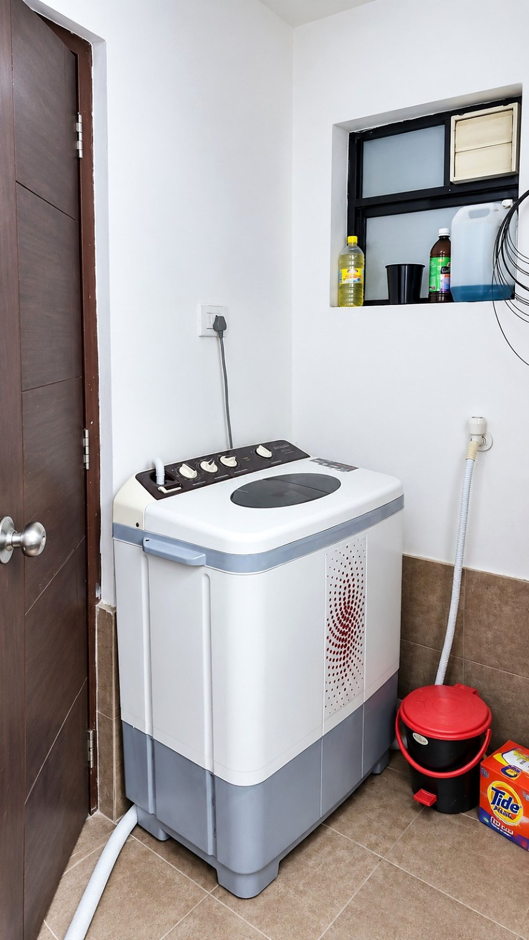 Une machine à laver compacte, de couleur blanche et grise, dotée d’un panneau de commande, d’un seau rouge et d’une étagère contenant des produits de nettoyage.