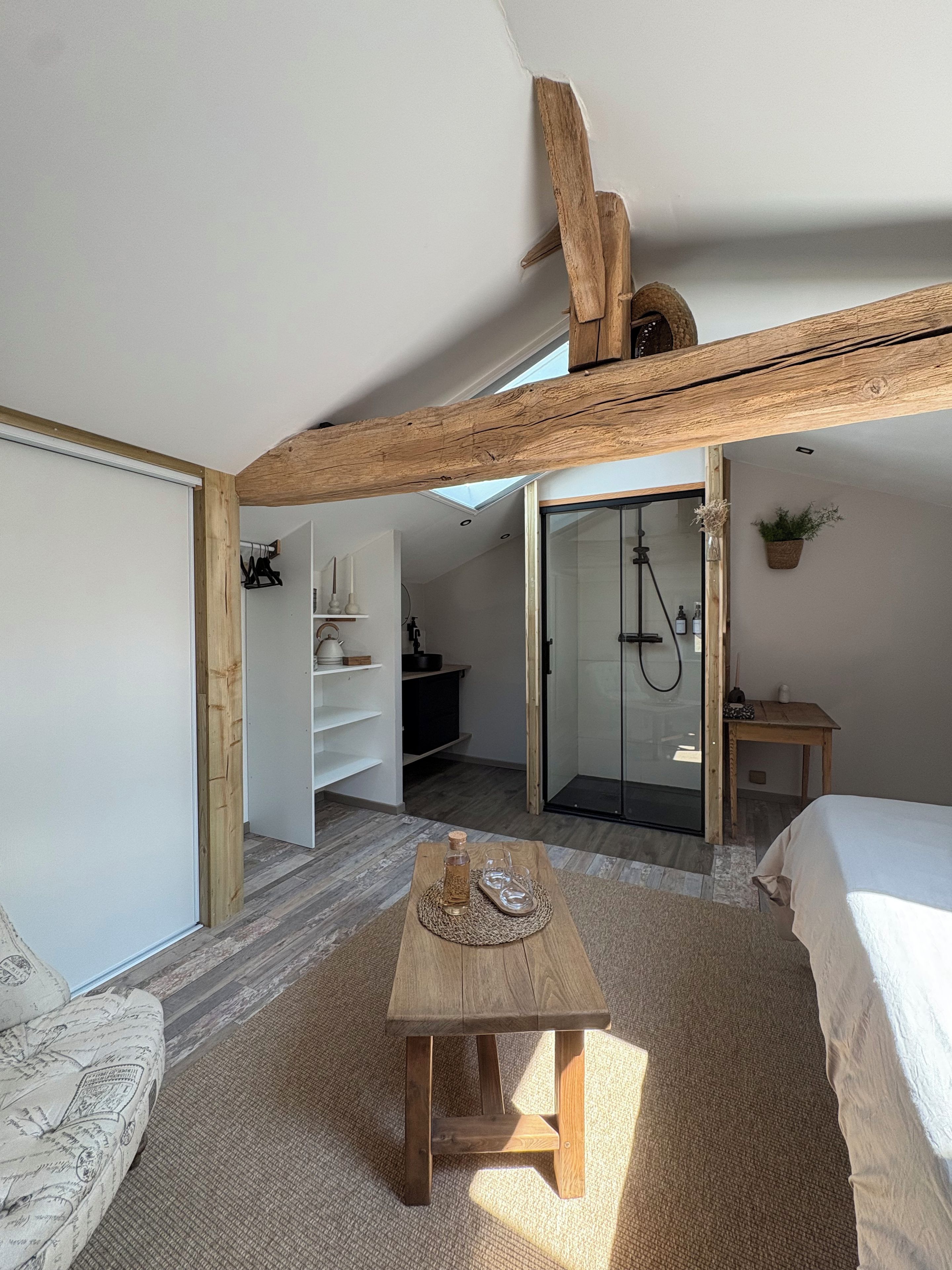 Een slaapkamer met een schuine kap, een houten tafel met een fles en glazen, een bed en een kleine tafel met een plant.