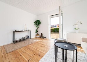 Una sala moderna con piso de madera, una mesa de centro negra, un sofá blanco, un ventanal que da al exterior y una planta pequeña en un estante.