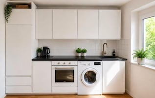 Cocina compacta con armarios blancos, encimera negra, horno integrado, lavadora y ventana con vistas.