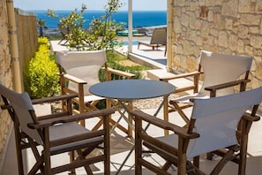 A wooden outdoor seating area with a round table, surrounded by chairs with white cushions, overlooking a coastal view.