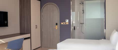 A hotel room with a bed, a desk, a chair, a wardrobe, and a shower area.