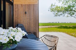 Ein Sitzbereich im Freien mit Tisch und Stühlen, ein Blumentopf mit weißen Blüten und Blick auf ein grünes Feld.