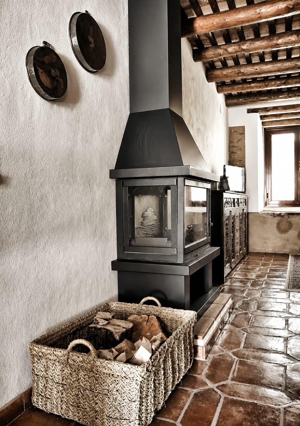 A stone-floored room with a fireplace, a woven basket of firewood, and a wooden ceiling.