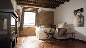 A room with a rustic wooden ceiling, a fireplace, a window with curtains, a sofa, a small table, and a wall-mounted artwork. - Tu refugio rústico con encanto andalusí: descanso, historia y naturaleza. (Constantina)