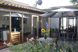 Een terras met een tafel en stoelen, een glazen deur naar een keuken, en een grote parasol die schaduw biedt.