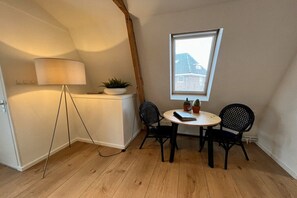 A small room with a wooden floor, a table with two chairs, a lamp, a window, and a plant on a white cabinet. - Achter de Duinen||Castricum (Castricum)