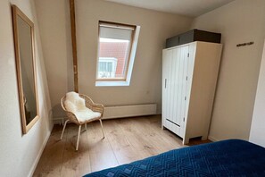 A room with a bed, a white wardrobe, a wicker chair, a radiator, and a window with a view of a building. - Achter de Duinen||Castricum (Castricum)