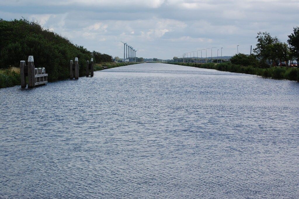 Un canal bordé d’une promenade en bois d’un côté et surmonté d’un pont métallique au loin.
