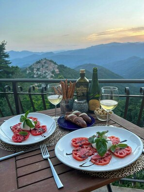 Een tafel gedekt met tomatensalade, broodstengels en flessen wijn, met uitzicht op een bergachtig landschap.