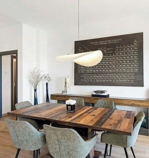 Ein Essbereich mit einem Holztisch, sechs Stühlen, einer Pendelleuchte und einem Wandbild des Periodensystems.