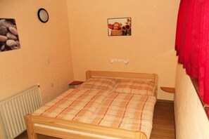 Ein Einzelbett mit kariertem Bettbezug in einem Zimmer, das einen Heizkörper, ein an der Wand befestigtes Bild und einen roten Vorhang zeigt.