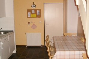 Eine kleine Küche mit Spüle, Heizkörper, einer Pinnwand mit Notizen und einem Essbereich mit Tisch und Stühlen.
