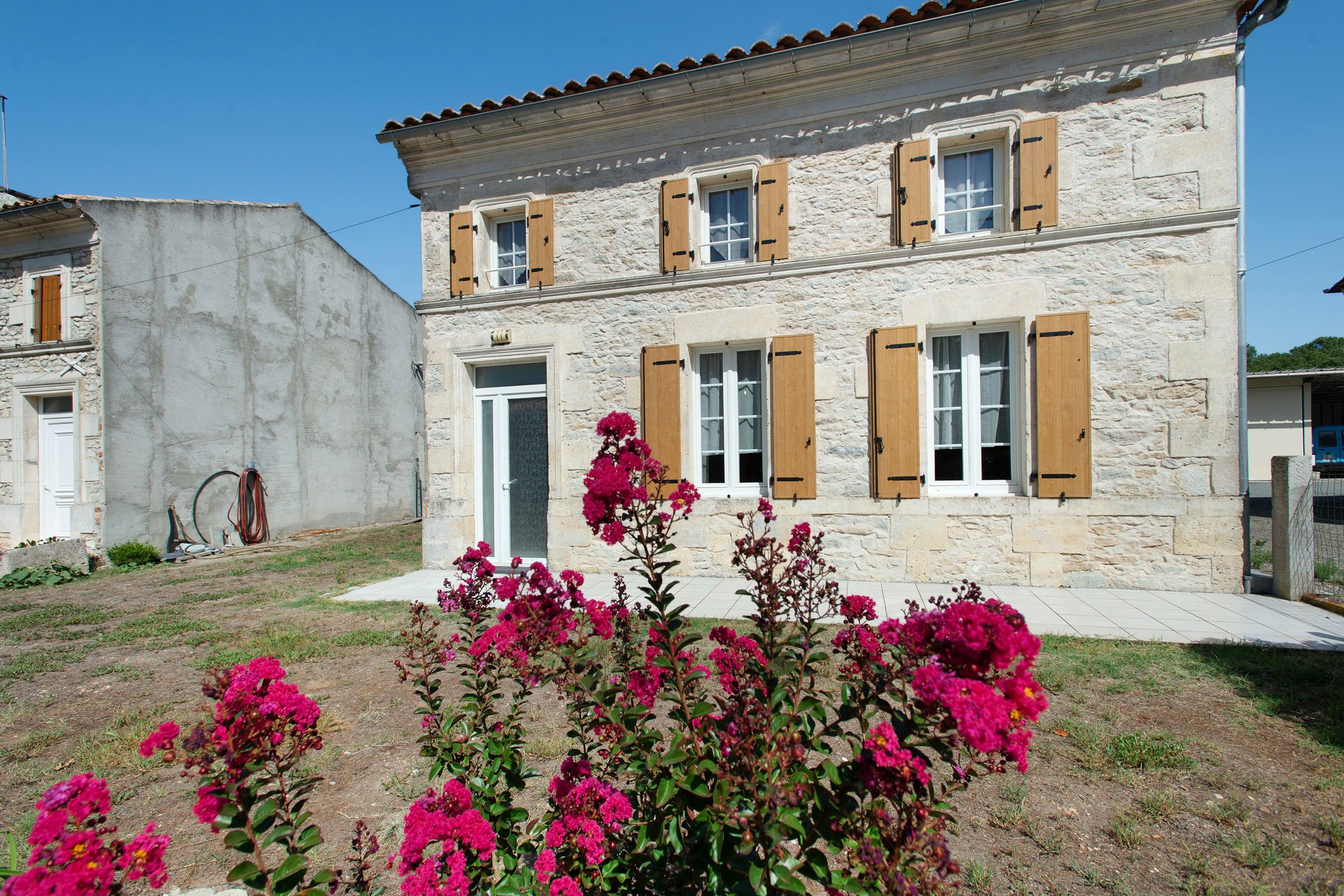 A two-story stone building with multiple windows and shutters, surrounded by a garden with pink flowers.