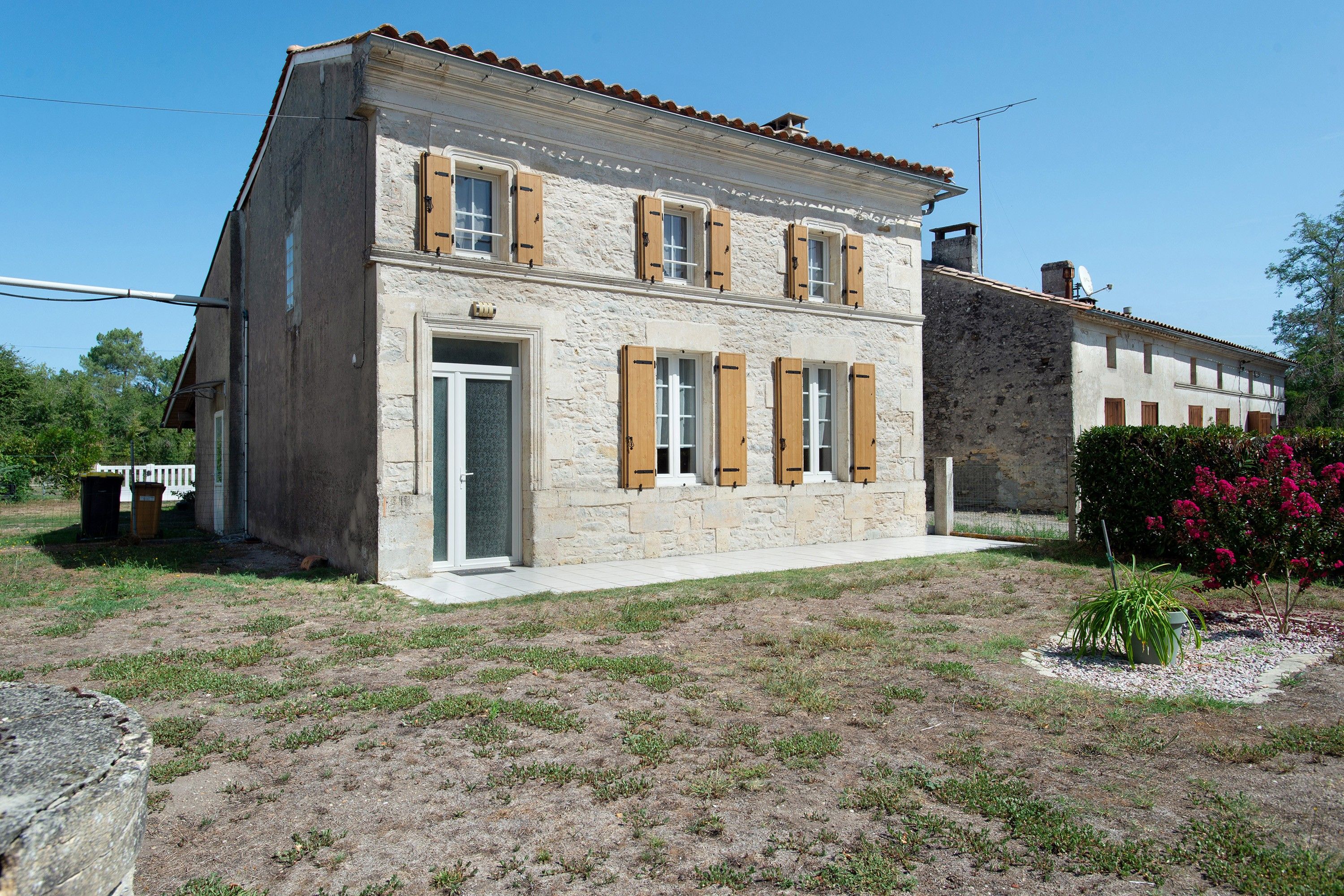A two-story stone building with a flat roof, wooden shutters, and a small garden with a potted plant.