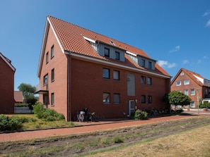 A red-brick residential building with a sloped roof, a visible garage door, and a small garden area with a bicycle. - Apartment 182, Sea Love (Juist)
