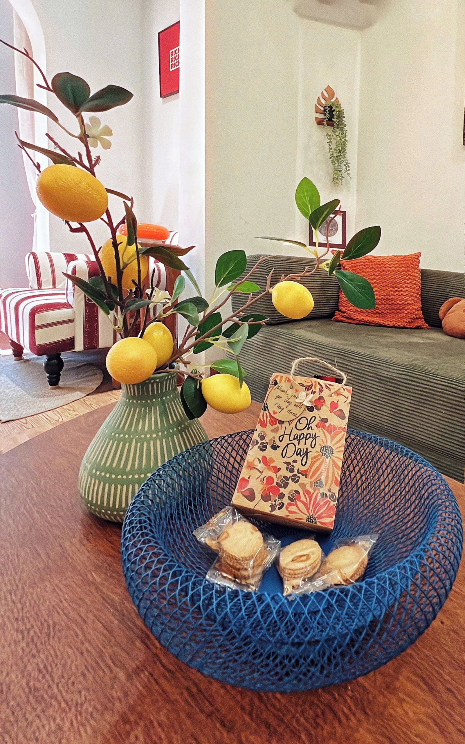 Un salón con un sofá a rayas, un jarrón verde con limones, una cesta azul con galletas y una tarjeta de saludo.