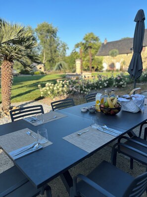 Una mesa de comedor al aire libre con un frutero, vasos y cubiertos, con un jardín y un edificio al fondo.