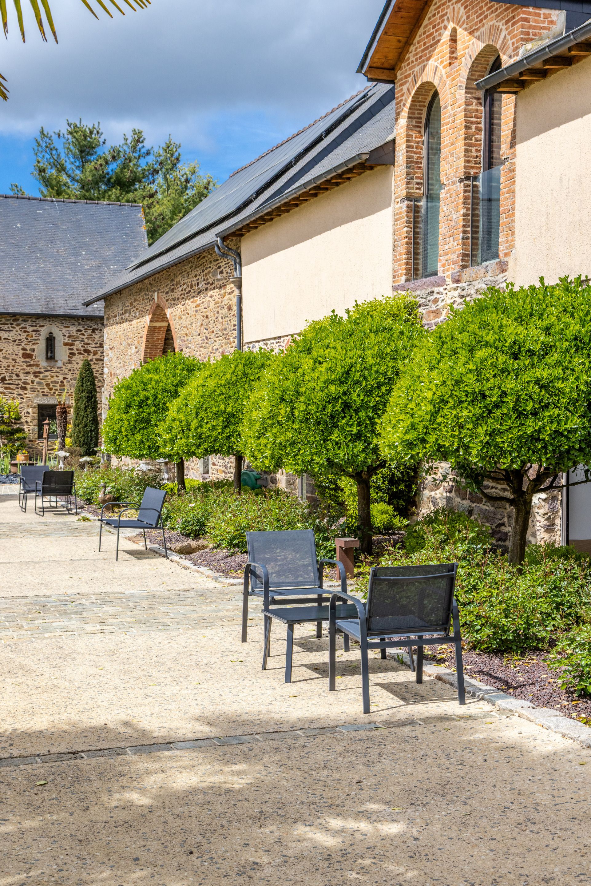 Une cour bordée de bâtiments en pierre, une zone pavée et plusieurs chaises.