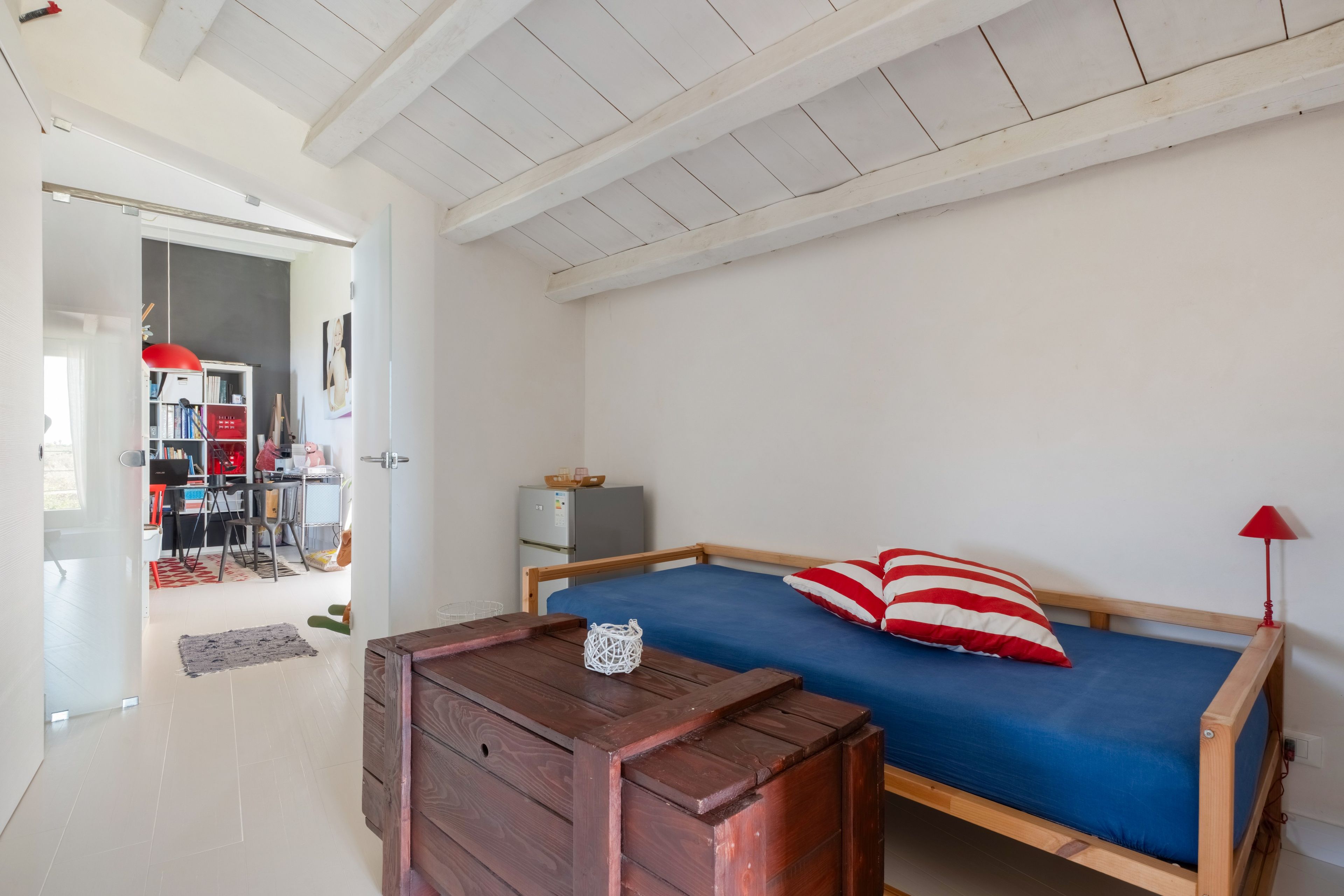 A room with a bed, a wooden table, a red lamp, and a white door leading to another room.