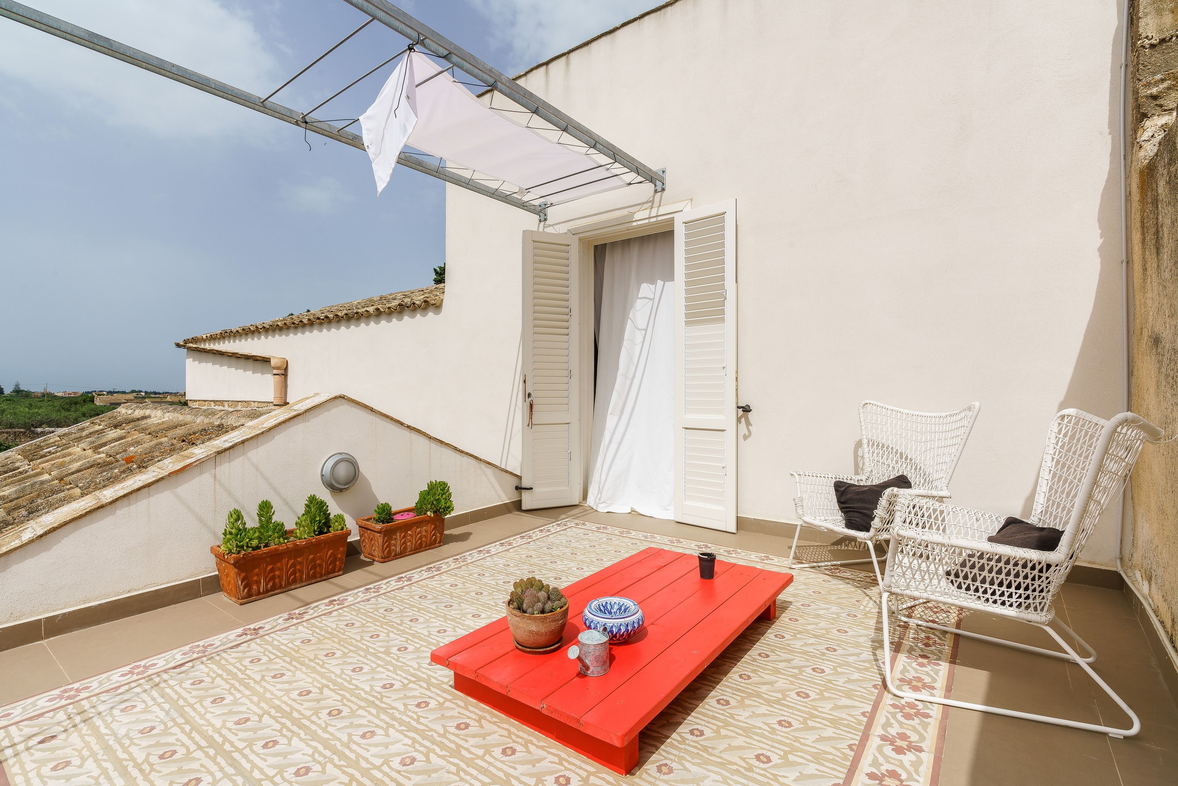 Une terrasse sur le toit avec une table rouge, deux chaises en osier blanches, des plantes en pot et un auvent blanc.