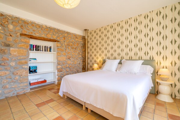 A bedroom with a stone wall, a bookshelf, a bed with white bedding, and patterned wallpaper. - Casa Barbarama - with private swimming pool (Alaro)