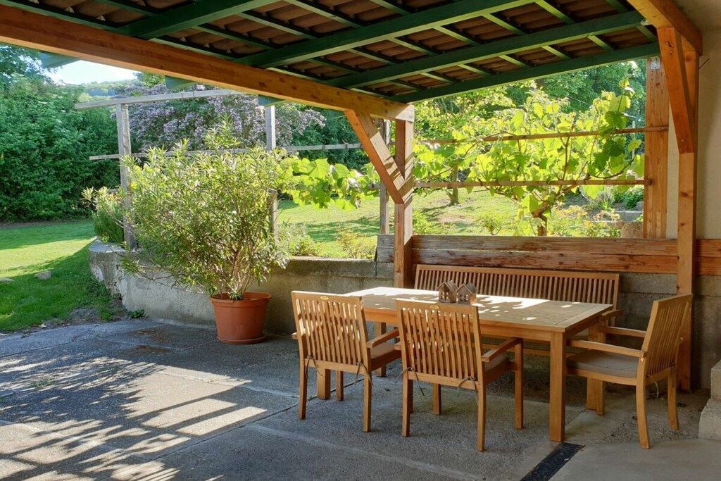 An outdoor wooden pergola with a table and chairs, surrounded by greenery and a garden.