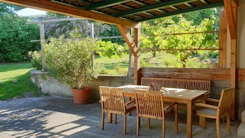 An outdoor wooden pergola with a table and chairs, surrounded by greenery and a garden.