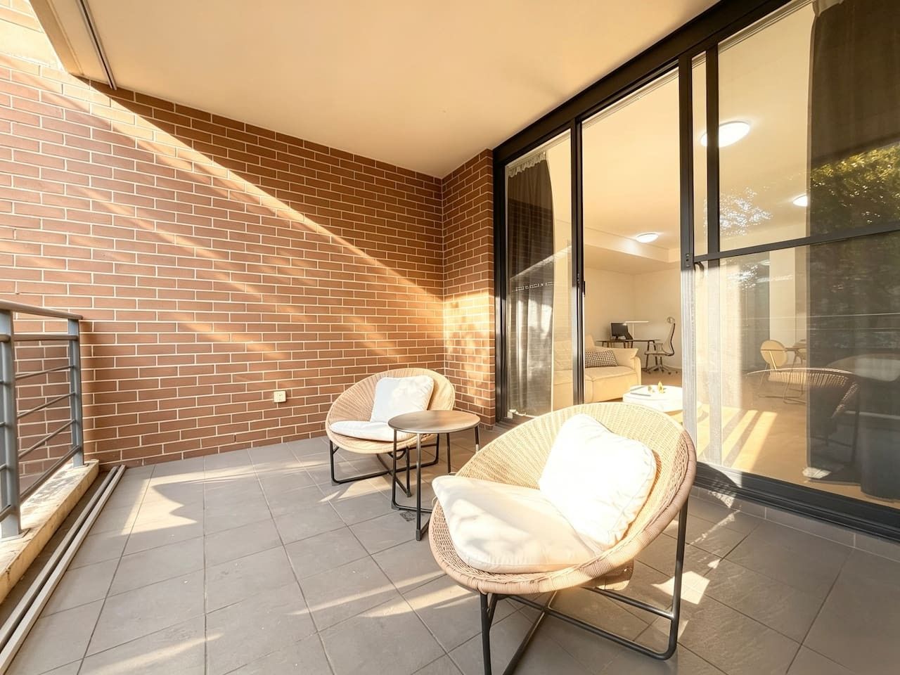 Eine sonnendurchflutete Terrasse mit zwei Korbstühlen und einem kleinen Tisch, einer Backsteinmauer und einer Glasdoor, die zu einem Außenbereich führt.