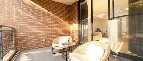 A sunlit patio with two wicker chairs and a small table, a brick wall, and a glass door leading to an outdoor area.