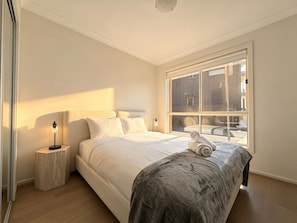 A bedroom with a large bed, a bedside lamp, a window with blinds, and a view of the outside. - JC House Park Side - 4 bedroom house (Bardia)