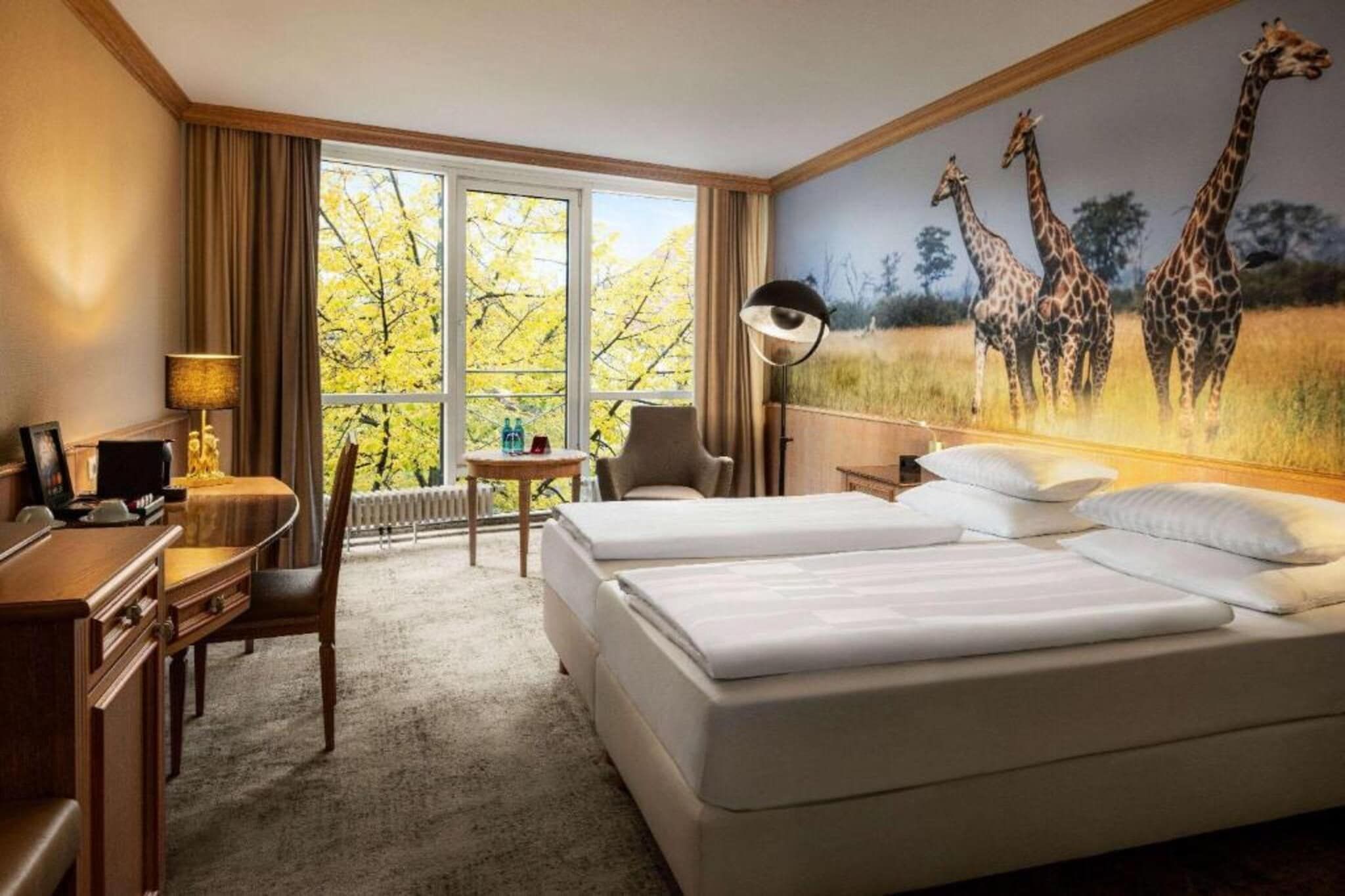 Een hotelkamer met twee bedden, een bureau, een stoel, een lamp en een groot raam met uitzicht op bomen.