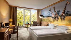 Et hotelværelse med to senge, et skrivebord, en stol, en lampe og et stort vindue med udsigt til træer.