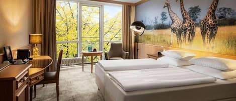 A hotel room with two beds, a desk, a chair, a lamp, and a large window with a view of trees.