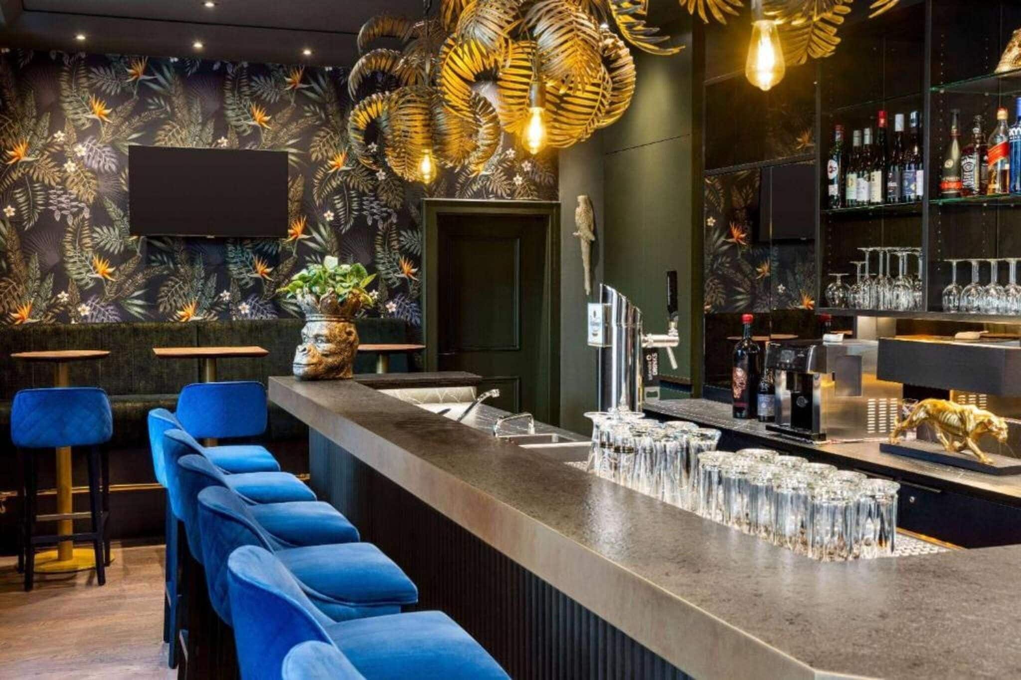 A bar area with a long counter, blue bar stools, a decorative plant, and a variety of bottles and glasses.